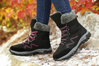 A person wearing black winter hiking boots with pink laces and gray fleece trim, standing on a snowy rock outdoors with autumn foliage in the background. The brand name OILAI is visible on the side of the boots.