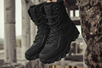 A pair of black winter combat boots with white faux-fur trim at the collar, held up by a person wearing camouflage pants in an outdoor urban setting.